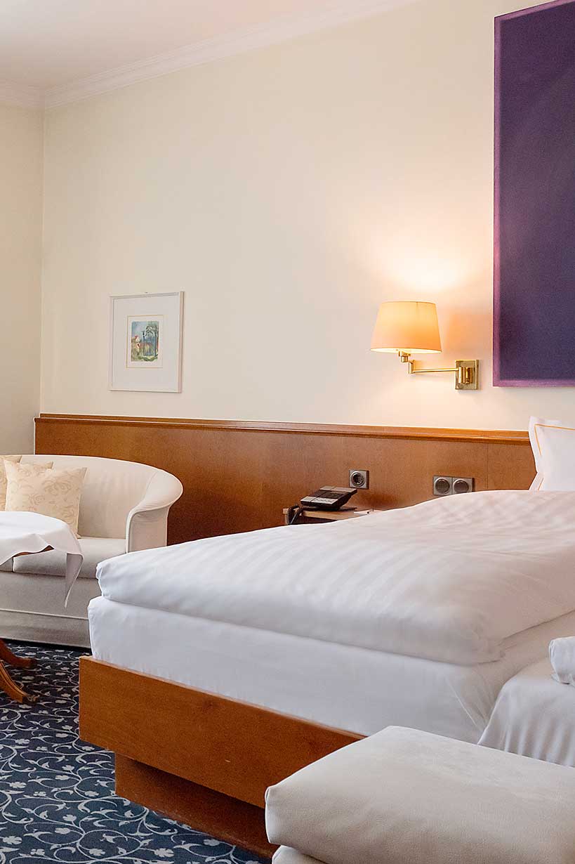 Ein ordentlich gemachtes Hotelbett neben einem weißen Sessel, mit einer Wandlampe und gerahmter Kunst über einem hölzernen Kopfteil – ideal für Ihren Aufenthalt in Fulda nach einer anstrengenden Konferenz.