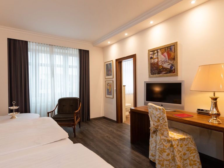Helles Hotelzimmer mit zwei Betten, einem Schreibtisch mit Fernseher und einem Fenster mit durchsichtigen Vorhängen und dunklen Gardinen.