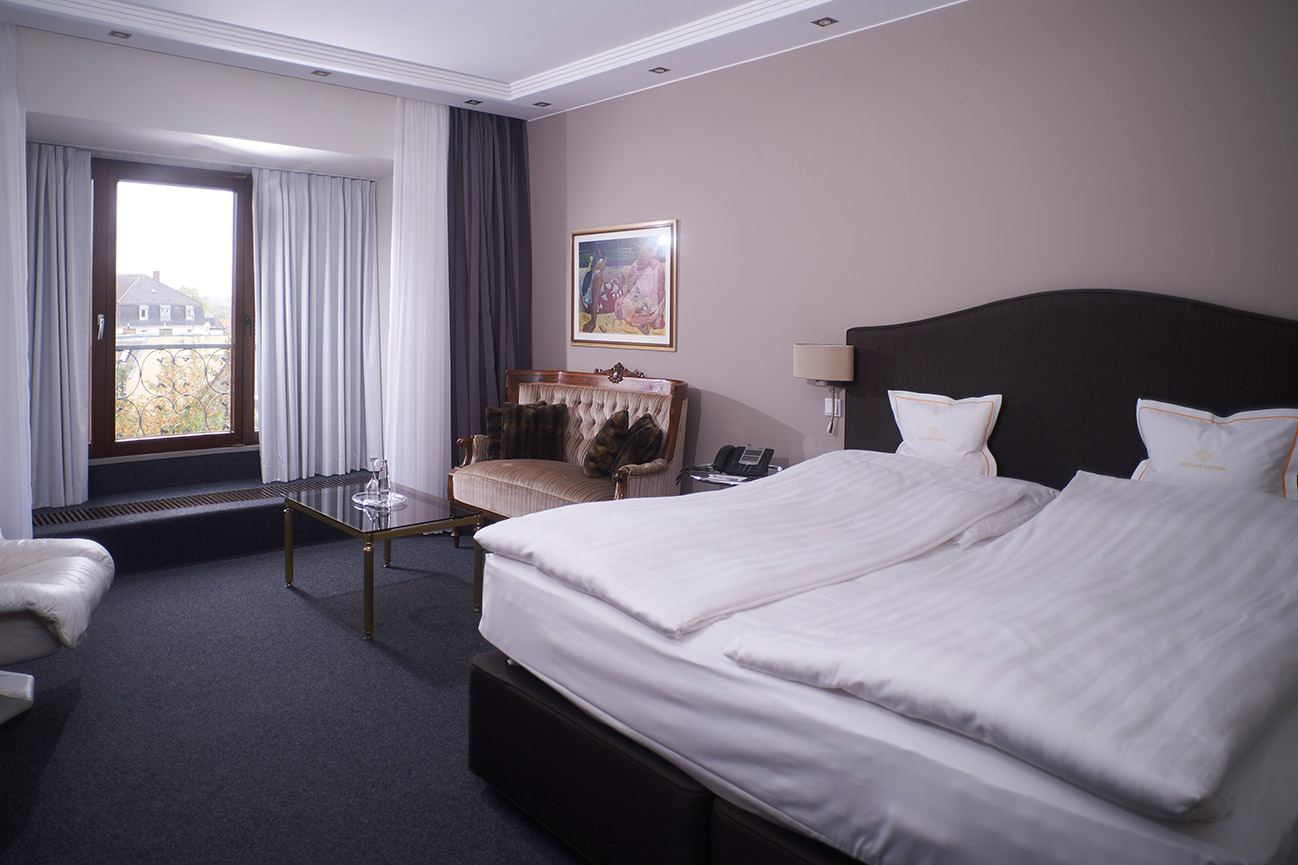 Hotelzimmer in Fulda mit zwei Betten, einem Sofa, einem Glastisch, einem großen Fenster mit Vorhängen und Wänden in neutralen Farben – perfekt zum Entspannen nach einer Konferenz.
