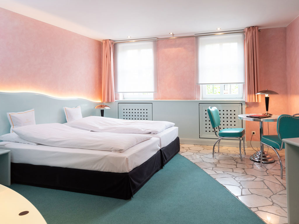 Goldener Karpfen Hotelzimmer mit pastellrosa Wänden, zwei Betten, türkisfarbenen Stühlen, einem Tisch und großen Fenstern mit weißen Jalousien – perfekt zum Entspannen nach einer Tagung.