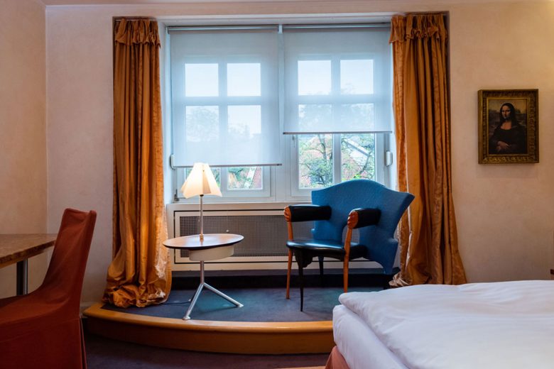 Ein Hotelzimmer in Fulda mit einem blauen Stuhl am Fenster, goldenen Vorhängen, einer Lampe und einem gerahmten Gemälde an der Wand – perfekt zum Entspannen nach Ihrer Tagung.