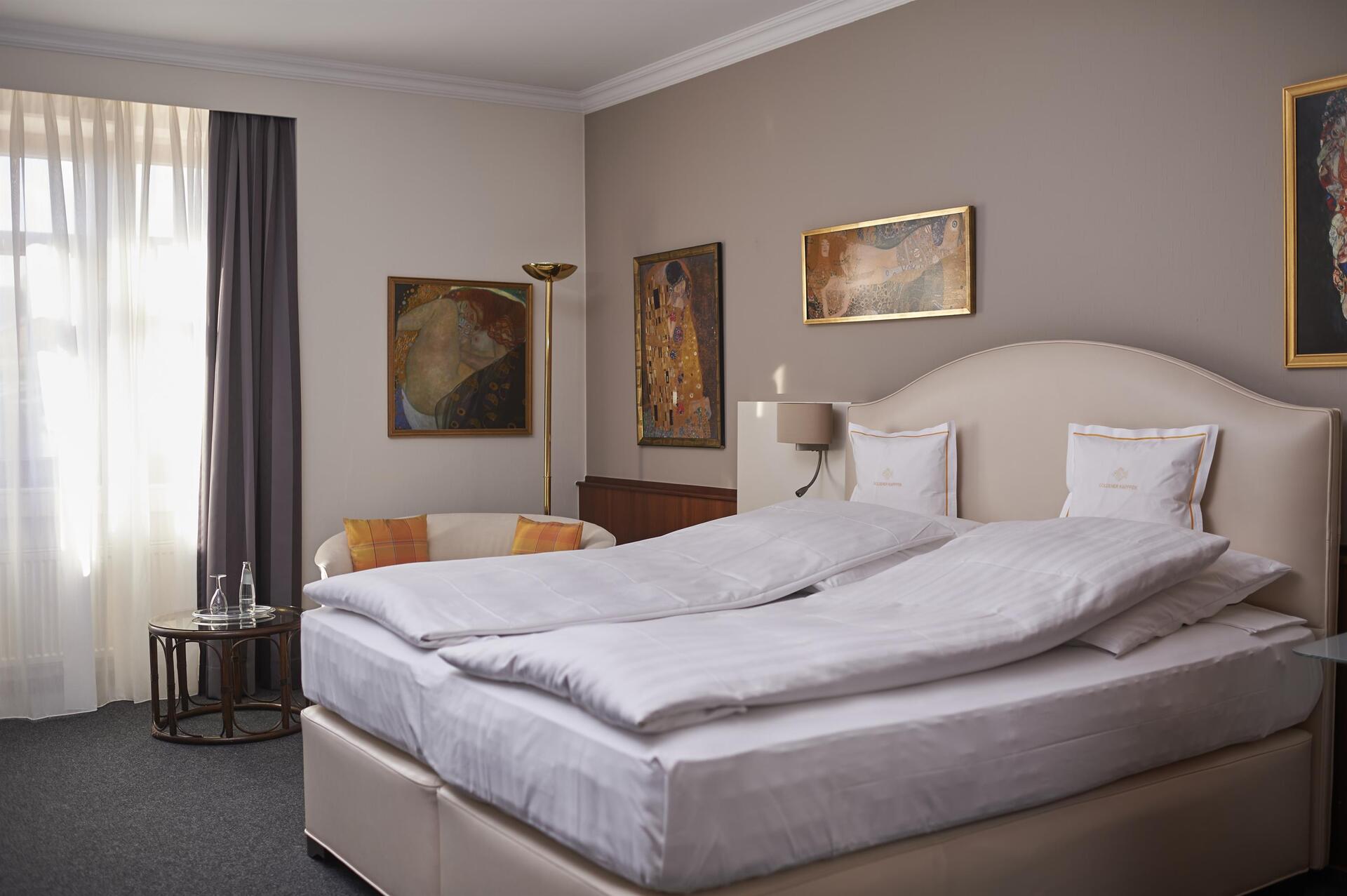 A neatly made bed offers classic comfort in a modern hotel room with art on the walls, a chair, and natural light streaming through the window.