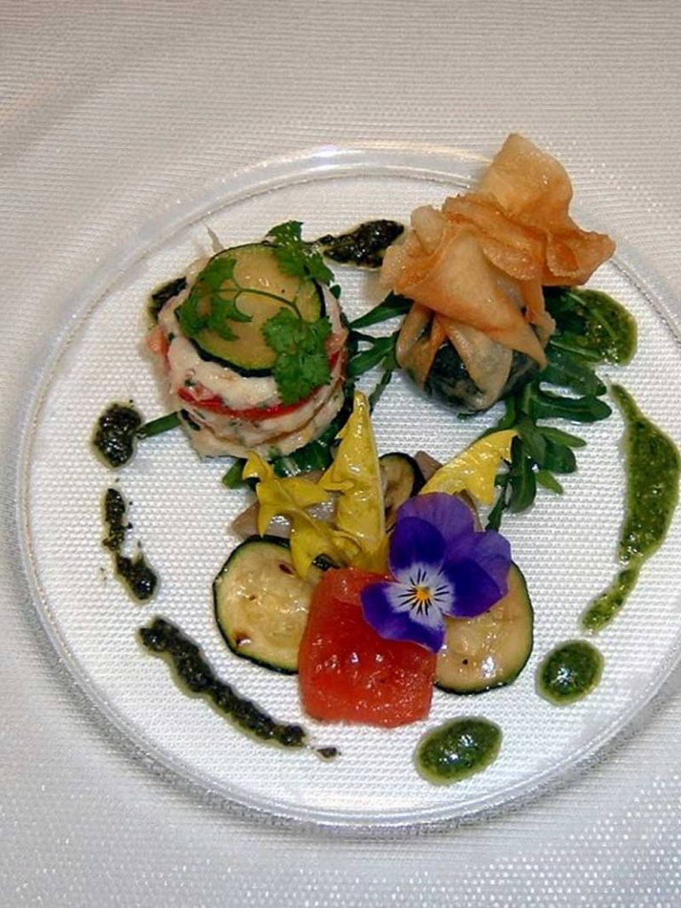 Ein Gourmet-Gericht in der Küche unseres Restaurants besteht aus Meeresfrüchten, Gemüse, Gebäck, Kräutern und einer essbaren lila Blume, die elegant auf einer klaren Glasplatte angerichtet ist.