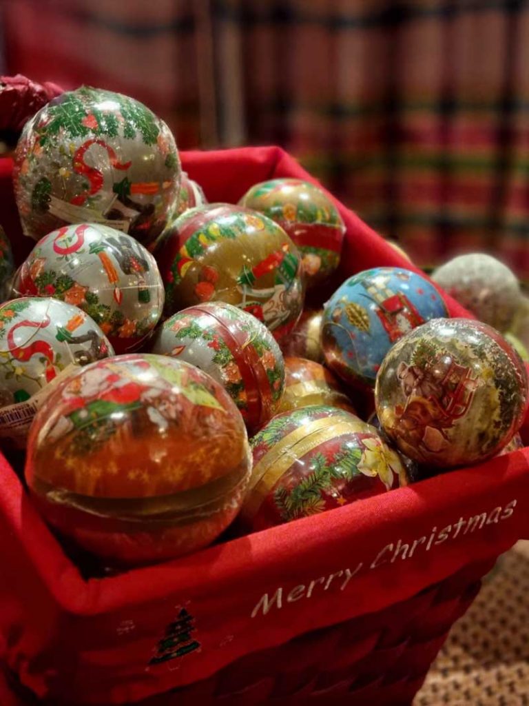 Roter Korb, gefüllt mit buntem Weihnachtsschmuck, mit gesticktem "Merry Christmas" an der Seite.