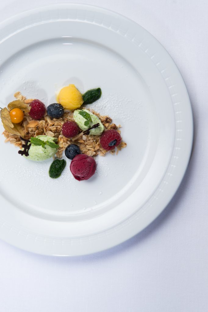 Ein weißer Teller mit verschiedenen Beeren, Eiskugeln und Müsli wird wunderschön auf einer weißen Tischdecke präsentiert und vermittelt die frische Eleganz einer guten Restaurantküche.