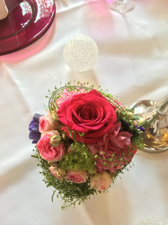 Kleiner Strauß mit einer großen roten Rose, rosafarbenen Blumen und Grünzeug, arrangiert auf einem weißen Tischtuch in einem Restaurant, neben einem Glasornament.
