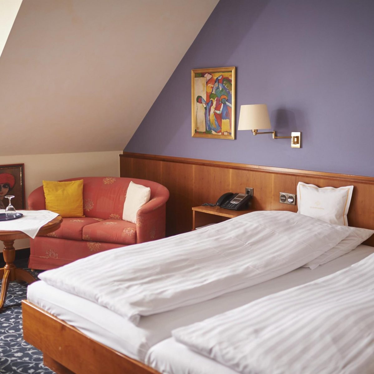 Gemütliches Hotelzimmer mit Doppelbett, Sofa, kleinem Tisch und bunten Kunstwerken an einer blauen Akzentwand.