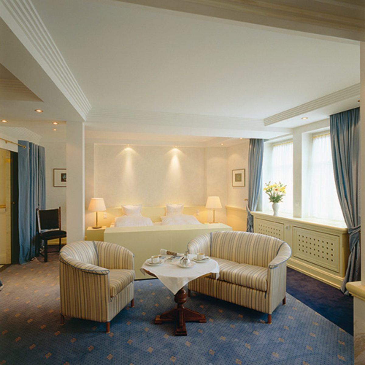 Elegantes Hotelzimmer mit zwei Einzelbetten, Sesseln, einem gedeckten Tisch für den Tee und großen Fenstern mit blauen Vorhängen.