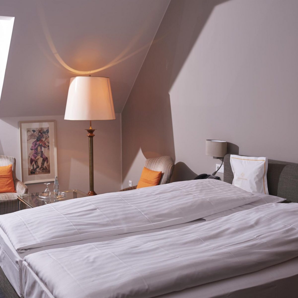 Hotelzimmer mit einem Doppelbett, zwei Sesseln, einer Stehlampe und einem Fenster mit durchsichtigen Vorhängen.
